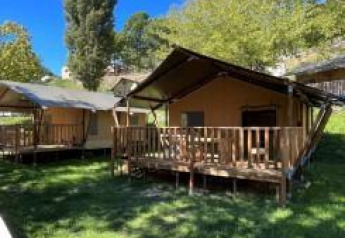 Safari tents with wooden decks set on green grass, surrounded by trees under a clear blue sky.