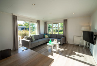 Salon lumineux avec canapé d’angle et grandes fenêtres dans un lodge Velthorst à Poort van Zeeland, Pays-Bas.