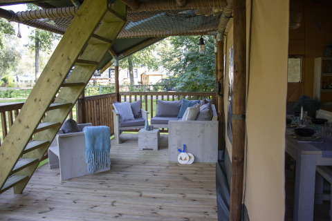 Terraza de madera con sofás y escalera en Safari tent Ranger en Camping Vossenberg, Países Bajos, rodeada de naturaleza.