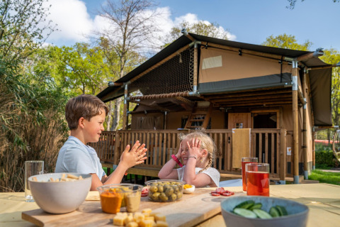Two children play at a table with snacks and drinks in front of a safari tent at Camping Vossenberg, Netherlands.