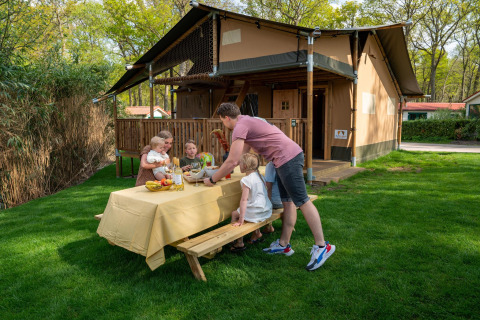 Familia disfrutando de una comida al aire libre junto a la tienda Safari Ranger en Camping Vossenberg, Países Bajos.
