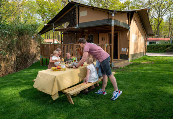 Familia disfrutando de una comida al aire libre junto a la tienda Safari Ranger en Camping Vossenberg, Países Bajos.