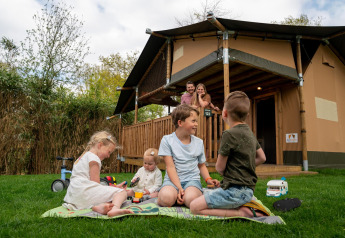 Niños juegan sobre el césped frente a una tienda safari en Camping Vossenberg, Países Bajos, con sus padres detrás.
