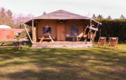 Tienda tipo safari Cottage en Camping Vossenberg en Países Bajos, con sillas y mesas de pícnic en el césped.