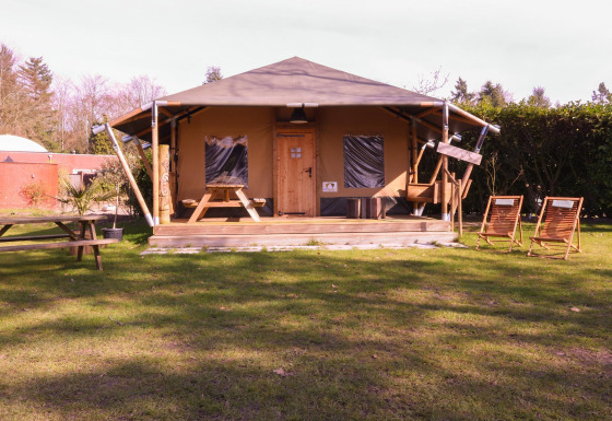Tienda tipo safari Cottage en Camping Vossenberg en Países Bajos, con sillas y mesas de pícnic en el césped.