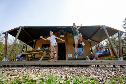Dos niños saltan de la veranda de una tienda safari en Camping Vossenberg, junto a una pareja y un perro.