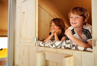Two happy kids relaxing on a loft bed in a Safari tent Cottage at Camping Vossenberg, Netherlands.