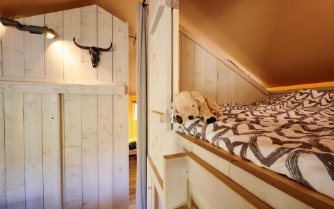 Vista interior de la Safari tent Cottage con cama elevada, peluche y decoración rústica en Camping Vossenberg.