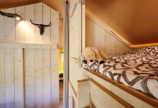 Vista interior de la Safari tent Cottage con cama elevada, peluche y decoración rústica en Camping Vossenberg.