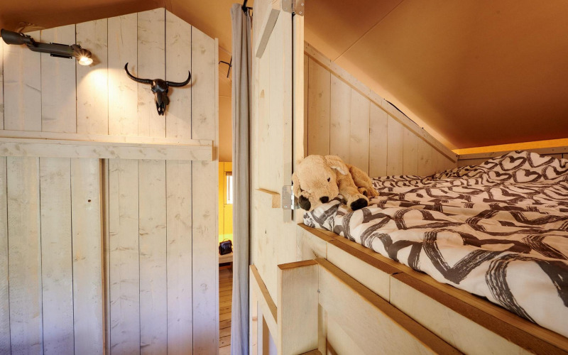 Vista interior de la Safari tent Cottage con cama elevada, peluche y decoración rústica en Camping Vossenberg.