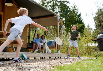 Dos niños juegan bádminton frente a una tienda safari en Camping Vossenberg, padres y perro observan.