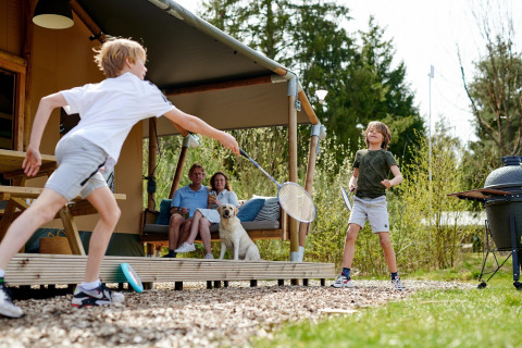 Deux enfants jouent au badminton devant une tente safari au Camping Vossenberg, parents et chien regardent.