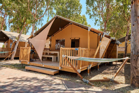 Tenda safari Villatent Outback con veranda in legno, amaca e alberi in un campeggio soleggiato.