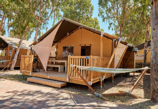 Tenda safari Villatent Outback con veranda in legno, amaca e alberi in un campeggio soleggiato.