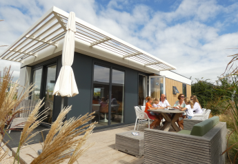 Familie sidder udenfor en moderne Dune Lodge ved Camping Zonneweelde i Holland under blå himmel.