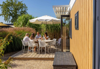 Family relaxing and dining outdoors at Dune Lodge Family, Camping Zonneweelde, Netherlands, under umbrella.