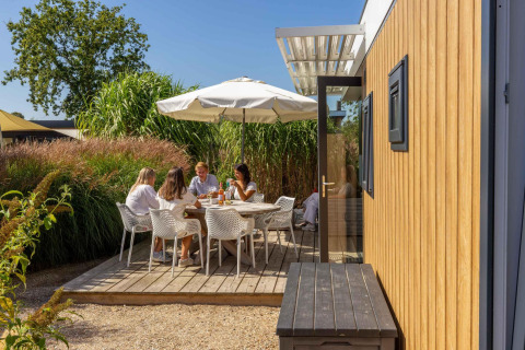 Family relaxing and dining outdoors at Dune Lodge Family, Camping Zonneweelde, Netherlands, under umbrella.