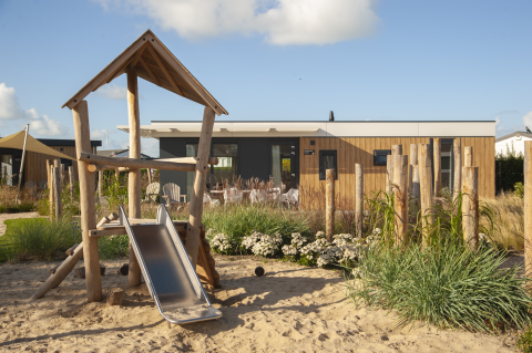 Spielplatz mit Rutsche vor einer Holzlodge bei Dune Lodge Family auf Camping Zonneweelde in den Niederlanden.