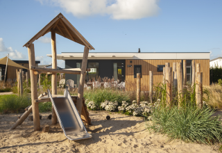 Spielplatz mit Rutsche vor einer Holzlodge bei Dune Lodge Family auf Camping Zonneweelde in den Niederlanden.