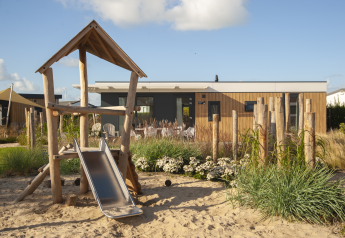 Speelplein met glijbaan voor een houten lodge bij Dune Lodge Family op Camping Zonneweelde, Nederland.