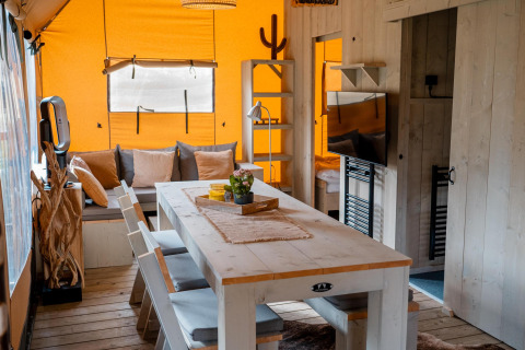 Vista interior de la tienda Safari Ranger DeLuxe con mesa comedor, sofá y decoración de madera en Camping Vossenberg.