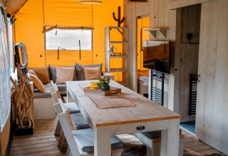 Vista interior de la tienda Safari Ranger DeLuxe con mesa comedor, sofá y decoración de madera en Camping Vossenberg.