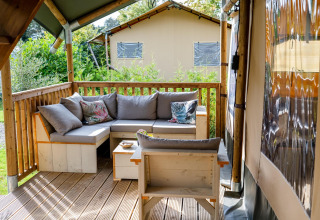 Zona salotto esterna con divano e cuscini su una veranda in legno presso una tenda safari al Camping Vossenberg.