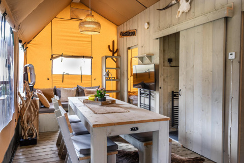 Interior de la tienda Safari Ranger DeLuxe con mesa de comedor, sofá y colores cálidos en Camping Vossenberg.