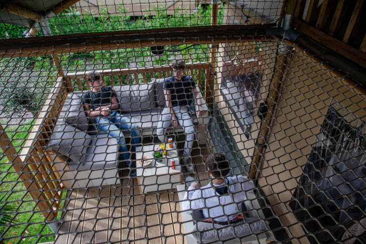 Tres personas se relajan en una terraza de una tienda safari en Camping Vossenberg, Países Bajos, vista a través de una red.