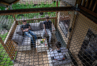 Tre persone si rilassano su una terrazza in una tenda safari al Camping Vossenberg, Paesi Bassi, viste attraverso una rete.