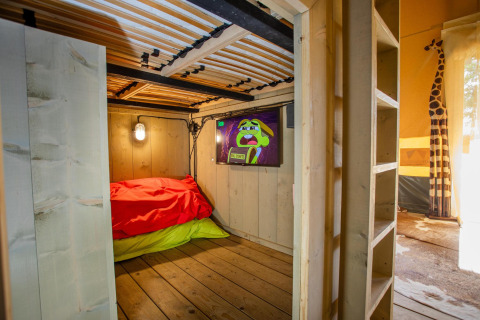 Vista interior de la tienda Safari Cottage Kaa en Camping Vossenberg, Países Bajos, con cama, TV y decoración.