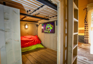 Vista interior de la tienda Safari Cottage Kaa en Camping Vossenberg, Países Bajos, con cama, TV y decoración.
