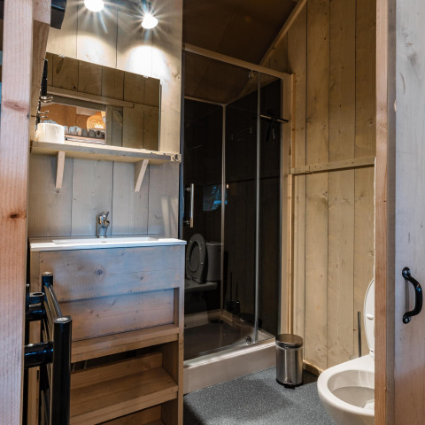 Baño en la tienda Safari Cottage Kaa en Camping Vossenberg, Países Bajos, con paredes de madera y ducha.