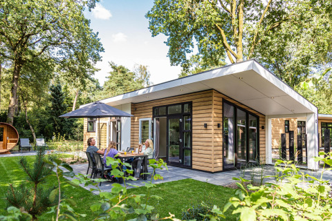 Cabaña moderna Silva sauna en entorno de bosque con familia disfrutando comida al aire libre bajo sombrilla.