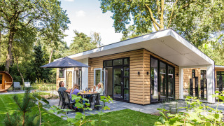 Cabaña moderna Silva sauna en entorno de bosque con familia disfrutando comida al aire libre bajo sombrilla.