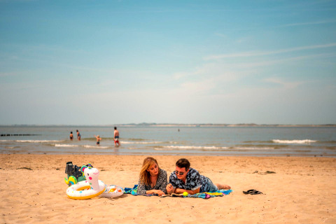 Ein Paar liegt auf einer Decke am Strand mit Schwimmutensilien, während Menschen im Meer baden.