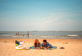 Un couple allongé sur une couverture à la plage avec des jouets, d'autres nagent en arrière-plan.