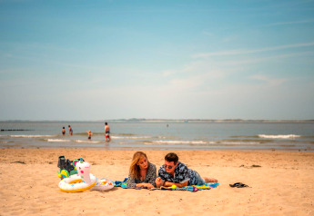 Et par ligger på et tæppe på stranden med strandudstyr, mens folk svømmer i havet i baggrunden.