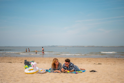 A couple relaxing on a beach blanket with swim gear, while others enjoy the sea in the background.