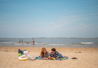 Et par ligger på et tæppe på stranden med strandudstyr, mens folk svømmer i havet i baggrunden.