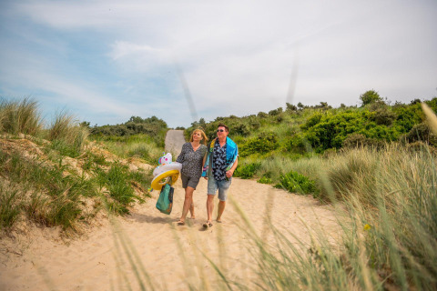 Un couple marche pieds nus dans les dunes près d’un lodge avec bouées et vêtements de plage.