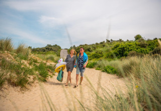 Una pareja camina descalza por las dunas de arena cerca de una cabaña, llevando flotadores y ropa de baño.