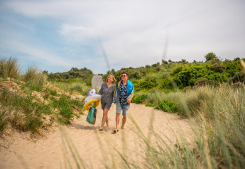 A couple walks barefoot through sandy dunes at a lodge, carrying swim gear and inflatable pool toys.
