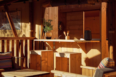Coin cuisine d’une tente safari avec mobilier en bois, plantes en pot, coussins et lumière du soleil.