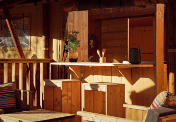 Zona cucina in una tenda safari con mobili in legno, piante, cuscini e calda luce del sole.