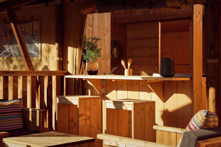 Cozy safari tent kitchen area featuring wooden furniture, potted plant, utensils, and cushions in the sun.