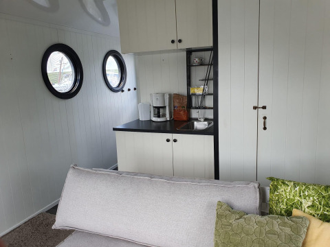 Lichte living en kitchenette met sofa, patrijspoorten en koffiemachine in een woonboot aan Rhederlaagse Meren.