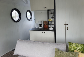 Sala luminosa con sofá, cocineta, ventanillas redondas y cafetera en una casa flotante en Rhederlaagse Meren.