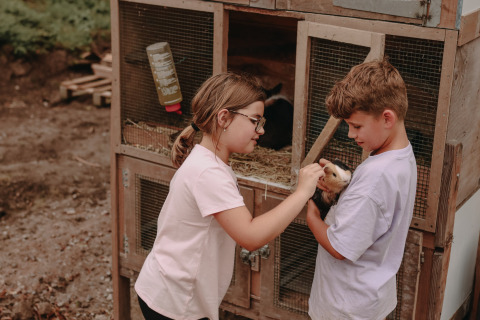 Zwei Kinder spielen mit einem Meerschweinchen vor einem Stall im Feather Down Fermette des trois tilleuls, Frankreich.