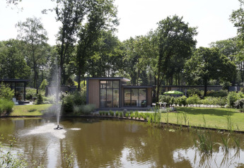 Uitzicht op een moderne lodge aan een vijver met fontein op Landgoed de Scheleberg in Nederland.